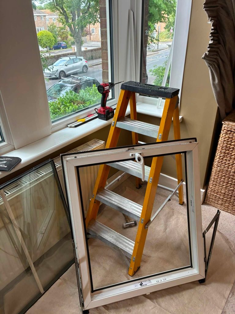 Removed uPVC window sash resting on a protective sheet next to a step ladder during hinge replacement.